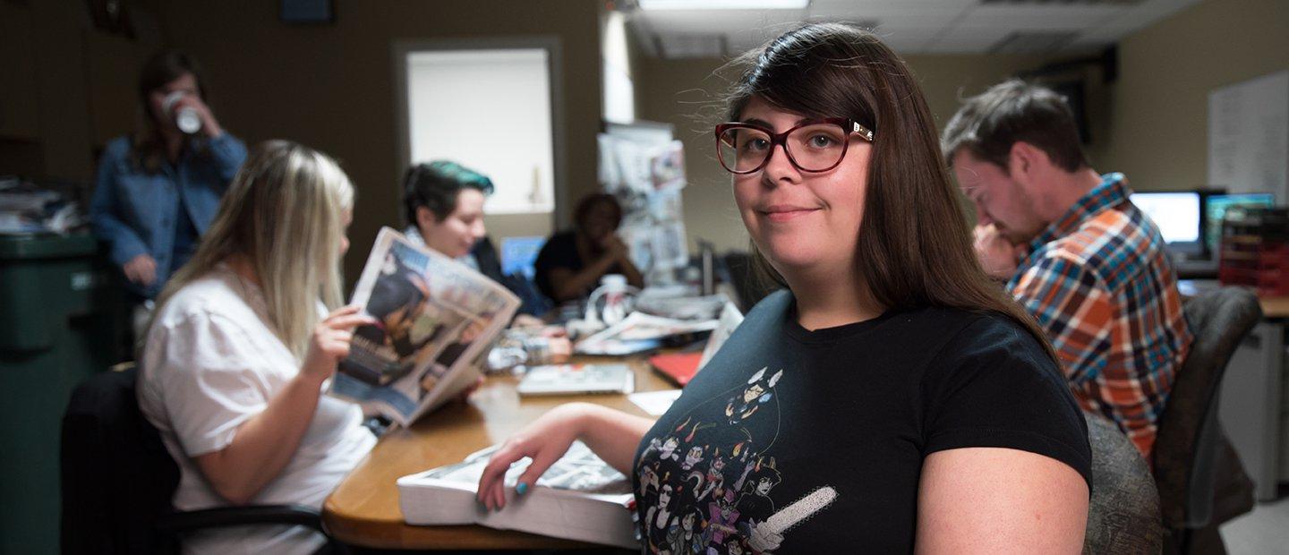 A woman faces the camera smiling. Her classmates are reading books and newspapers in the background